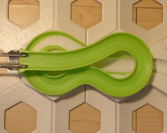 Pode incluir: Uma peça de pista verde de plástico em forma de infinito com conectores de metal, parte de uma pista de mármore ou brinquedo de construção semelhante. A pista está colocada sobre um fundo de azulejos hexagonais brancos e de madeira.