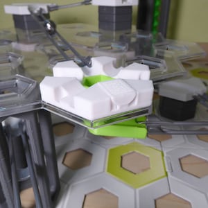 Puede incluir: Primer plano de una pieza de recorrido de canicas de plástico blanco con una palanca de plástico verde. La pieza está unida a una pista de plástico transparente y a una pista de metal gris.