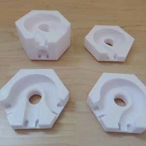 Könnte beinhalten: Vier weiße Kunststoffformen in Hexagonform mit einem gekrümmten Kanal in der Mitte. Die Formen sind unterschiedlich groß.