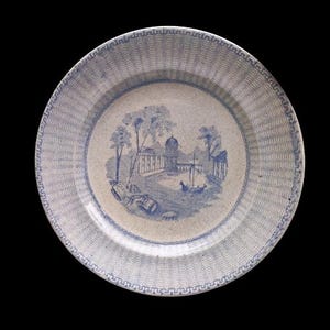 Può includere: Un piatto in ceramica blu e bianco con un bordo decorativo e un'immagine centrale di un edificio con alberi e una barca. Il piatto è in buone condizioni, senza scheggiature o crepe visibili.