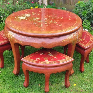 Puede incluir: Una mesa redonda lacada en rojo y marrón con tres taburetes a juego. La mesa y los taburetes tienen diseños florales dorados. La mesa está rodeada de hierba verde y plantas.