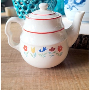 May include: White ceramic teapot with a lid and a curved handle. The teapot is decorated with red stripes and colorful floral designs. The teapot is a charming addition to any kitchen.