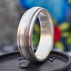 Peut inclure: Une bague en argent avec un design rainuré. La bague est posée sur un fond flou de fleurs. La bague porte les initiales "VJ" gravées à l'intérieur. La bague est posée sur une surface noire.