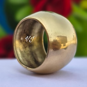 May include: A gold-colored, cylindrical ring with rounded edges. The ring has a smooth, polished surface and a hollow interior. The initials "VJ" are visible on the side of the ring. The background is blurred with hints of blue, red, and green.