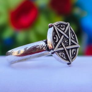 May include: Silver-toned ring featuring a pentagram design within a circular frame. The ring has a polished finish and a detailed, engraved pattern. The background is blurred, with hints of green, red, and blue.