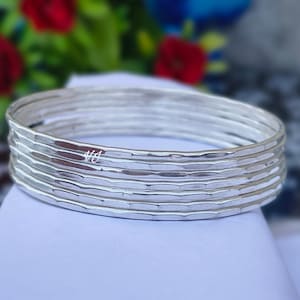 May include: A set of five stacked silver bangles, each featuring a hammered, textured surface. The bracelets are circular and metallic, with a small, faint logo reading VJ visible on the surface of the second bangle from the top.