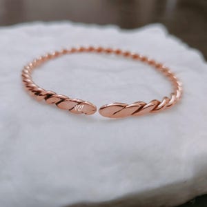 Könnte beinhalten: Ein roségoldenes, gedrehtes Armkettchen. Das Armband ist offen und hat ein schlichtes Design.