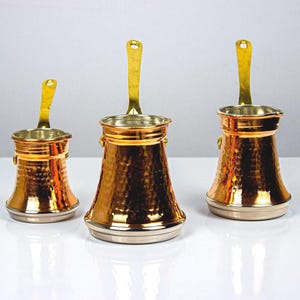 Puede incluir: Tres cafeteras de cobre con textura martillada y asas doradas. Las cafeteras tienen un diseño clásico con una base ancha y una parte superior estrecha. Las asas son curvas y se extienden hacia arriba. Las cafeteras están dispuestas sobre una superficie blanca.