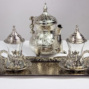 May include: Ornate silver tea set with a teapot, two glass cups, and a serving tray. The teapot and cups have intricate designs. The set is displayed on a white surface.