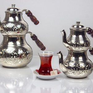 Puede incluir: Dos teteras plateadas ornamentadas con asas de madera oscura y patrones florales decorativos. Un vaso de té y un plato pequeño con un terrón de azúcar están frente a las teteras. Las teteras están apiladas con una base bulbosa y una parte superior más pequeña.