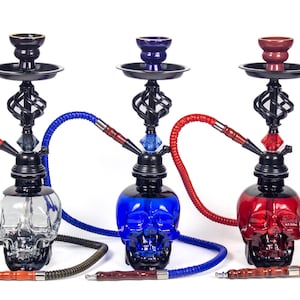 May include: Three skull-shaped hookahs in clear, blue, and red glass. Each hookah has a black metal stem with a decorative swirl design, a bowl, and a colored hose. The hoses are black, blue, and red.