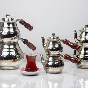 Puede incluir: Una colección de teteras turcas plateadas y brillantes con asas de madera. Las teteras están dispuestas con un vaso de té y un plato pequeño. El té es de color rojo intenso y las teteras tienen una textura martillada.