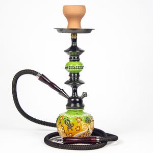 Puede incluir: Una cachimba decorativa con un tallo y una manguera negros, con una base estampada en verde y amarillo. La cachimba tiene un cuenco de arcilla marrón y una bandeja de metal negro. El diseño incluye patrones florales y geométricos.
