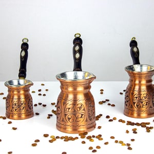 以下が含まれることがあります： 装飾的なデザインと濃い木製のハンドルが付いた、3つの銅製のコーヒーポット。各ポットは銀色の内側と小さな注ぎ口が付いています。コーヒー豆がポットの周りに散らばっています。