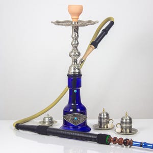 Puede incluir: Una cachimba azul con detalles plateados, una manguera beige y un cuenco de arcilla. Dos pequeñas tazas y platillos plateados están en primer plano. La cachimba tiene un diseño decorativo en la base azul.