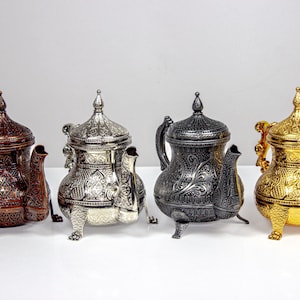 May include: Four ornate teapots in a row, each with a unique metallic finish. The teapots are brown, silver, black, and gold, all featuring intricate engraved patterns, spouts, and handles. They stand on small, decorative feet.