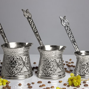 Könnte beinhalten: Drei silberfarbene türkische Kaffeekannen in verschiedenen Größen, jede mit einem verzierten Blumendesign und einem langen Griff, der von einer Tulpenform gekrönt wird. Die Kannen werden auf einer weißen Oberfläche mit Kaffeebohnen und gelben Blumen präsentiert.