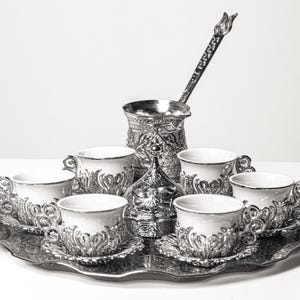 Puede incluir: Un juego de café plateado con seis tazas y platillos, una cafetera, un azucarero y una cuchara de mango largo, todo sobre una bandeja decorativa. Las tazas y la cafetera tienen diseños florales ornamentados.