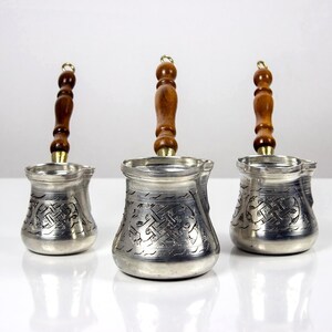 Puede incluir: Tres cafeteras plateadas con mangos de madera. Cada cafetera tiene un diseño grabado y un mango largo y curvo de madera. Las cafeteras están dispuestas en fila, con los mangos apuntando hacia arriba.