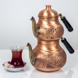 Puede incluir: Una tetera de dos niveles de color cobre, con diseños grabados ornamentados y asas negras. Un vaso de té y un plato pequeño con delicias turcas están en primer plano. La tetera tiene tapa y boquilla con detalles decorativos.