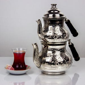 Könnte beinhalten: Eine silberfarbene, zweistöckige Teekanne mit aufwendigen geprägten Mustern, schwarzen Griffen und einem passenden Deckel. Ein Glas roter Tee und ein kleiner Teller mit türkischem Honig ergänzen die Präsentation. Das Teeservice steht auf einer weißen Oberfläche.