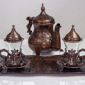 May include: An ornate copper-colored tea set. The set includes a teapot, two glass cups with decorative holders, and saucers, all resting on a matching tray. The pieces feature intricate embossed patterns.