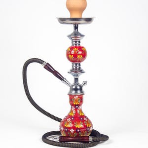 Puede incluir: Una cachimba roja con diseños florales amarillos y detalles plateados. Tiene una manguera oscura y un cuenco de arcilla. La base y el cuerpo central están adornados con diseños detallados. La cachimba está diseñada para fumar.