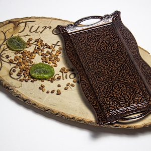 May include: A decorative wooden serving tray with a dark brown, ornate, rectangular tray on top. The tray is placed on a rustic, oval wooden platter with text and dried fruit and seeds.