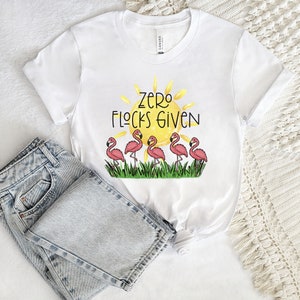 Puede incluir: Camiseta blanca con un sol amarillo y flamencos rosas. El texto "Zero Flocks Given" está impreso en la camiseta.