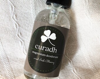May include: A clear glass bottle with a black spray top and a brown label. The label has a green shamrock and the text "curadh magnesium deodorant a real Irish Blessing".