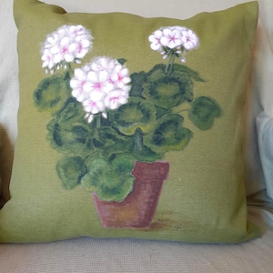 May include: A square cushion with a painted floral design. The cushion has a green background with a potted plant with white and pink flowers. The pot is brown, and the leaves are green.