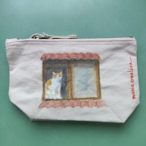 Pode incluir: Uma bolsa de lona bege com zíper com uma ilustração pintada de um gato em uma janela. O gato é branco e laranja. A janela tem um telhado vermelho. O texto "mania creativa" é impresso no lado direito.
