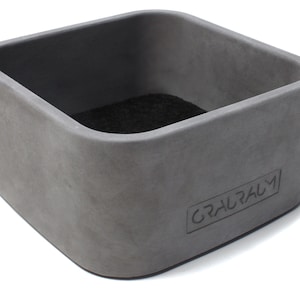 Könnte beinhalten: Ein quadratischer Betontopf in Grau mit einem schwarzen Filzeinsatz und dem eingeprägten Wort "GRALRALM" auf dem Boden.