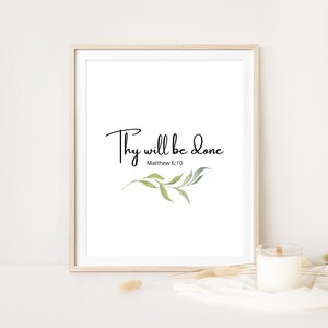 Puede incluir: Una impresión enmarcada con el texto "Thy will be done" y "Matthew 6:10" en escritura negra. La impresión presenta una ilustración de acuarela de hojas verdes y se muestra sobre un fondo blanco. Una vela y objetos decorativos están en primer plano.