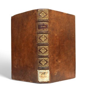 May include: A vintage book with a worn, brown leather cover. The spine features gold decorative elements and the title "CANTIQUE DES CANTIQUE" in gold lettering. The book has a weathered appearance, suggesting age and historical significance.