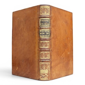 May include: A tan leather-bound book with a spine featuring gold-tooled floral patterns and two black labels with gold lettering that reads NOUVE TESTAM and TOM XI. The book is standing upright, showing the front cover and the detailed spine.