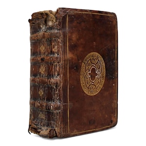 Può includere: Un libro antico, rilegato in pelle marrone scuro con dettagli dorati. Il dorso presenta nervature e elementi decorativi. Un emblema dorato adorna la copertina. I bordi del libro mostrano segni di usura.