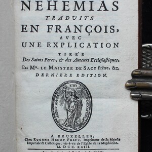 May include: A vintage book cover with the title "ESDRAS ET NEHEMIAS TRADUITS EN FRAN&Ccedil;OIS, AVEC UNE EXPLICATION TIREE Des Saints Peres, & des Auteurs Eccleſiaſtiques. Par MR. LE MAISTRE DE SACY Pr&ecirc;tre, &c. DERNIERE EDITION." The book was published in Brussels by Eugene Henry Frick in 1723.