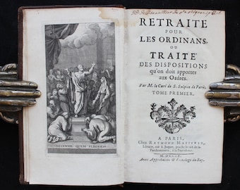 May include: An open antique book with a detailed black and white illustration on the left page and French text on the right. The illustration depicts a religious scene with figures. The book is held open by a decorative metal book holder.
