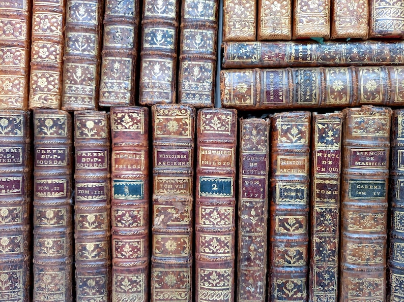 May include: A collection of antique books with aged, textured leather covers. The spines feature gold embossed floral and geometric designs, with titles and volume numbers in various fonts. The books are arranged closely together, creating a visually rich display.
