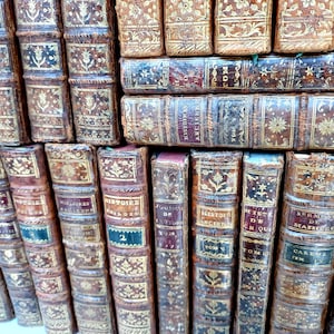 May include: A collection of antique books with aged, brown leather covers. The spines feature gold lettering and decorative patterns. Some books have labels with titles and volume numbers. The books are stacked together.