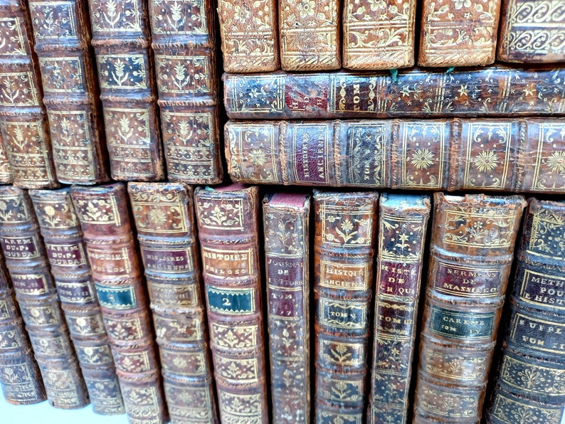 May include: A collection of antique books with aged, brown leather covers. The spines feature gold lettering and decorative patterns. Some books have labels with titles and volume numbers. The books are stacked together.