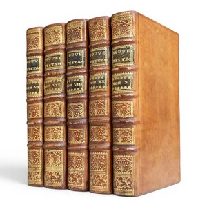 May include: Five antique books with tan leather covers standing upright. The spines feature gold-tooled floral patterns and black labels with the text NOUVEAU TESTAM and volume numbers ranging from VI to X in gold lettering on each individual spine.