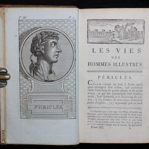 以下が含まれることがあります： 「LES VIES DES HOMMES ILLUSTRES」というタイトルと「PERICLES」という名前の開いた本が表示されています。左ページには、月桂樹の冠をかぶった男性の肖像画が描かれています。この本はフランス語で書かれており、詳細なテキストが記載されています。