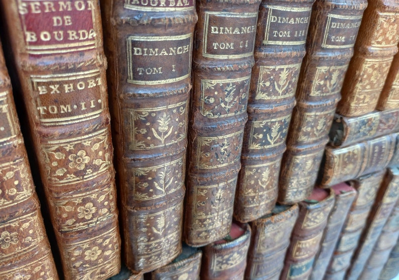 May include: A collection of antique books with brown leather covers and gold lettering and decorative accents. The spines display titles like "Dimanch" and "Sermon de Bourdaloue". The books are arranged in a stacked formation.