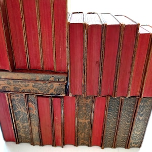 May include: A stack of antique books with red covers and gold accents. The spines are visible, showing the titles and the aged texture of the paper. Some books have patterned covers in shades of brown and gray.