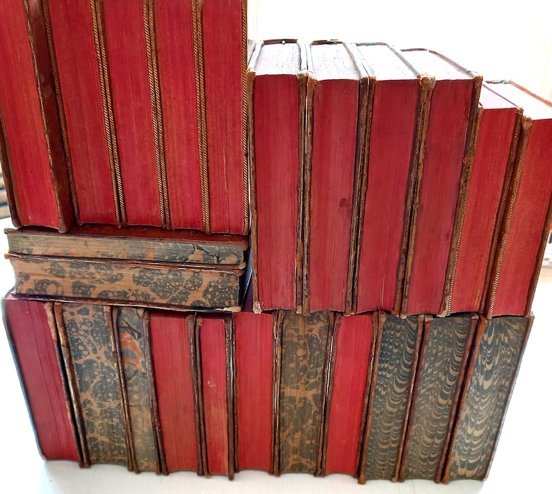 May include: A stack of antique books with red covers and gold accents. The spines are visible, showing the titles and the aged texture of the paper. Some books have patterned covers in shades of brown and gray.