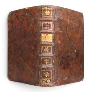 May include: An antique leather-bound book with a mottled brown cover and a raised spine. The spine features gold-tooled decorative panels and a dark red label with the text SECRE DE MEDEC. The leather shows signs of wear and surface scuffing.