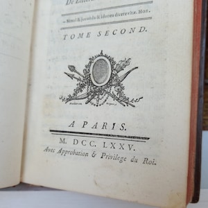 May include: Open book page with text and an ornate emblem. The text reads "TOME SECOND" and "A PARIS. M. DCC. LXXV." The emblem features a central oval surrounded by arrows, a torch, and floral elements. The book's edges are red.