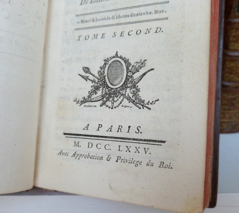 May include: Open book page with text and an ornate emblem. The text reads "TOME SECOND" and "A PARIS. M. DCC. LXXV." The emblem features a central oval surrounded by arrows, a torch, and floral elements. The book's edges are red.